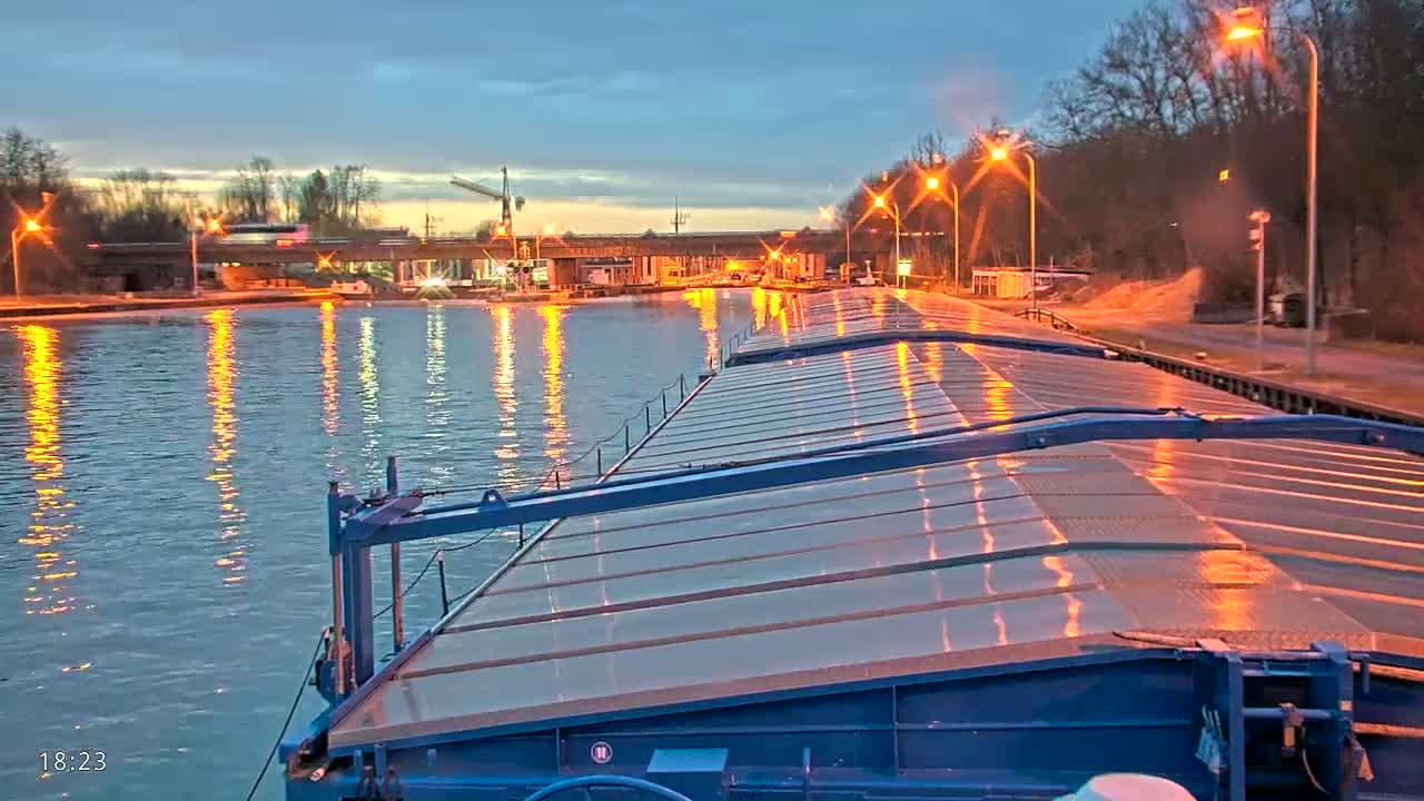 A barge on a calm waterway at dusk, illuminated by lights along the banks.