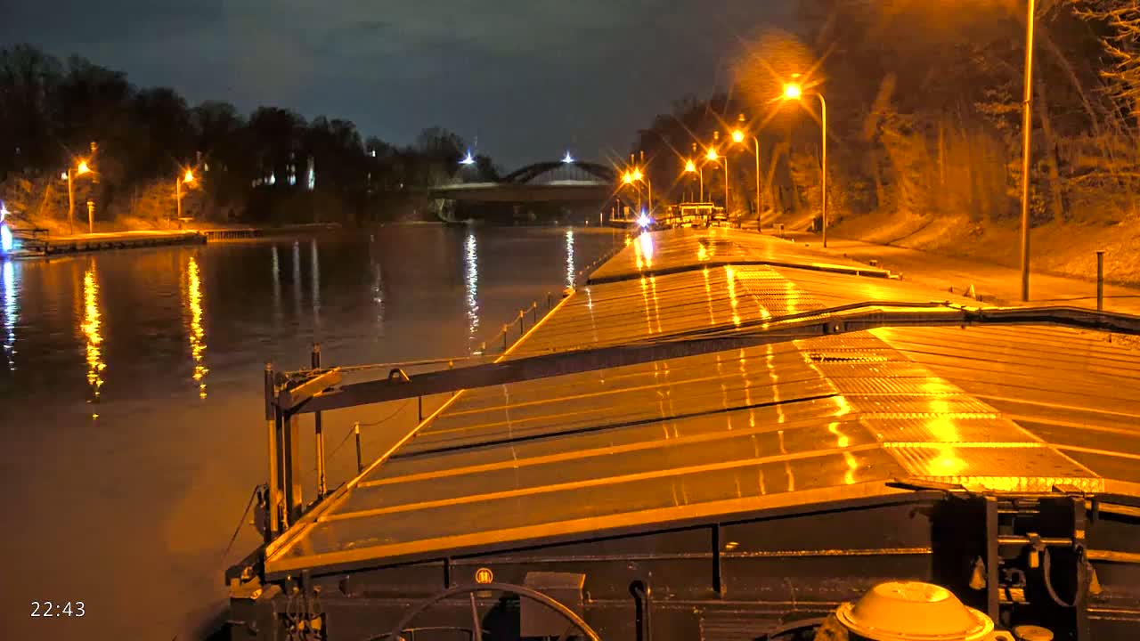 A barge's wet deck is illuminated at night by streetlights lining a canal, with a bridge visible in the distance.