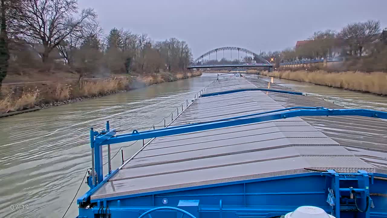 A barge with a blue deck travels down a river under an overcast sky, passing under a bridge in the distance.