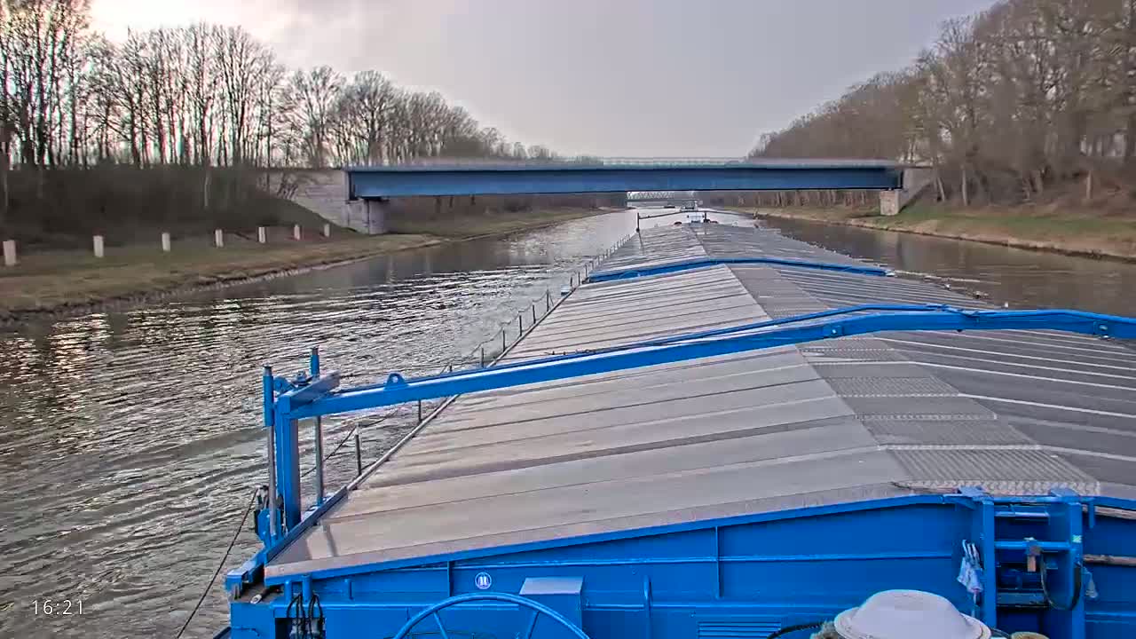 A blue barge travels down a canal under an overcast sky, passing under a bridge lined with bare trees.