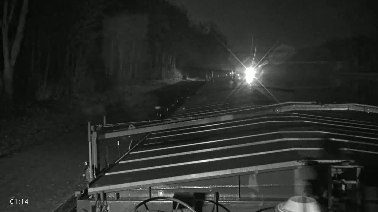A nighttime view from the driver's seat of a paving machine shows a dark road ahead with headlights visible in the distance.