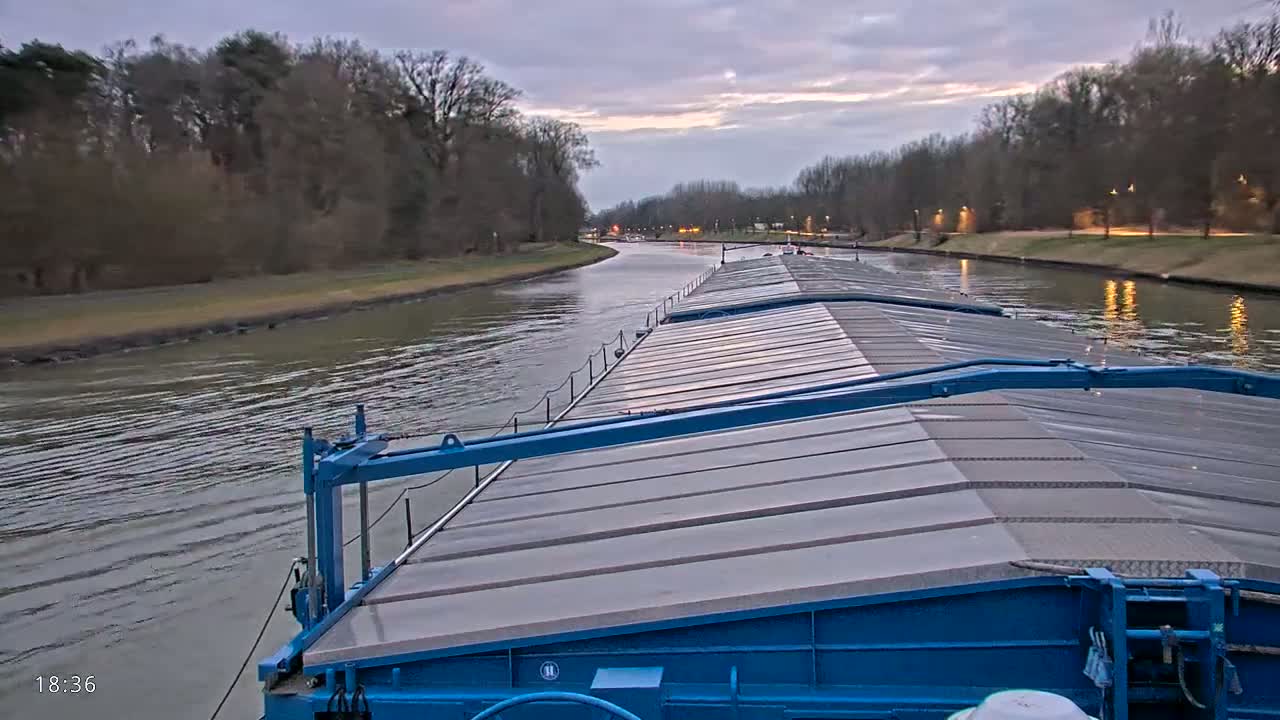 A blue barge travels down a canal at dusk, under a cloudy sky, past tree-lined banks with lights visible in the distance.