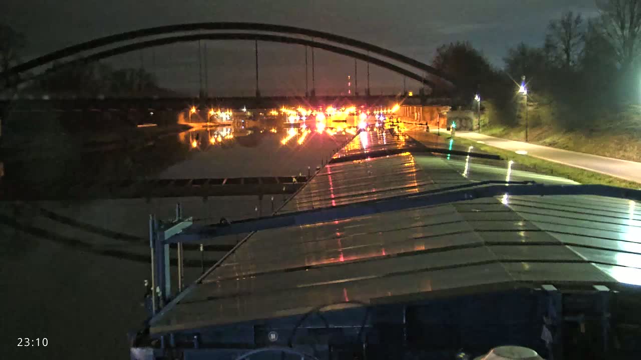 A nighttime view from a barge on a canal, showing a double-arched bridge in the distance illuminated by lights, reflecting on the water's surface.