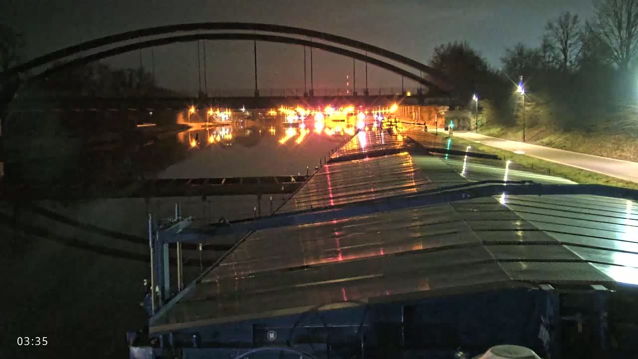 At night, a barge travels along a canal under a large arched bridge, with lights reflecting on the water's surface.