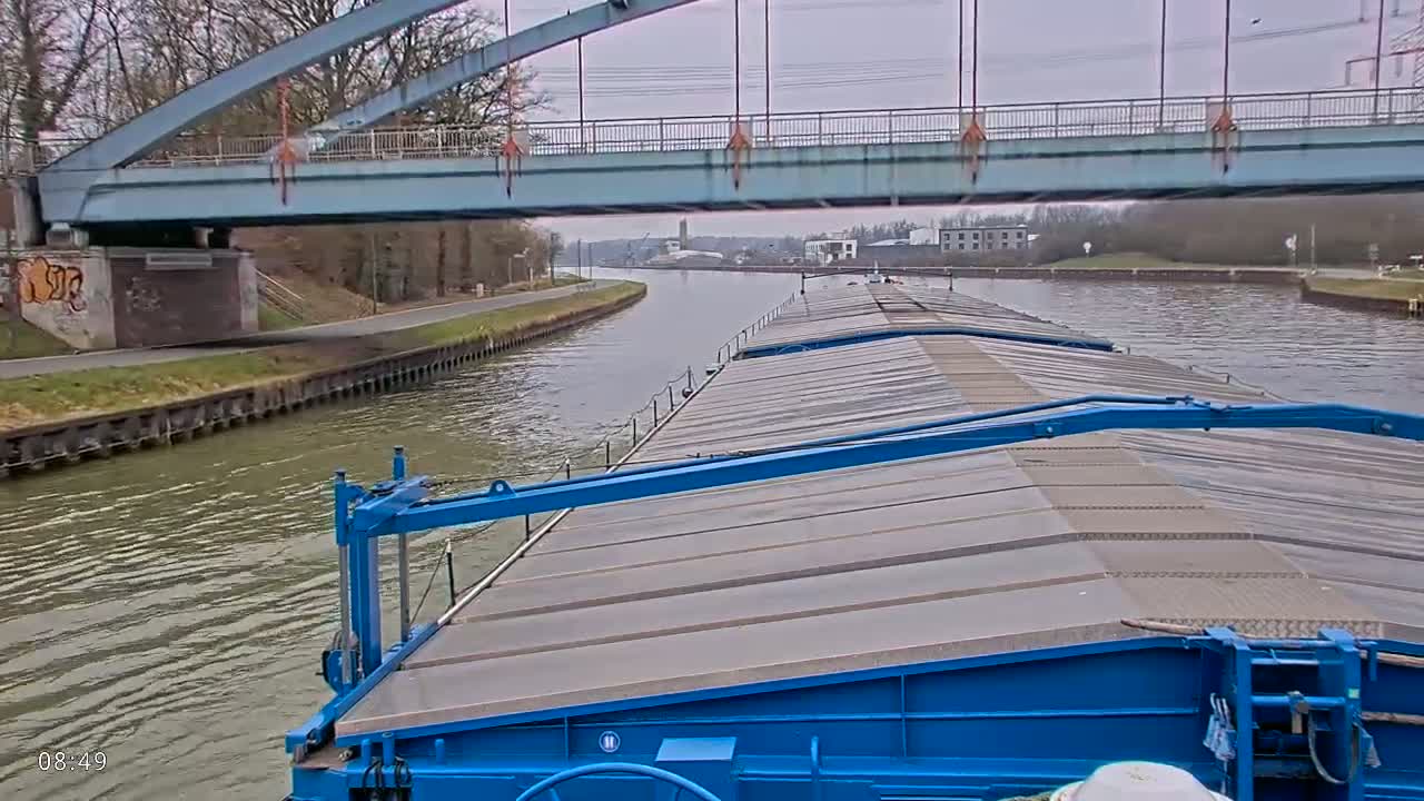 A blue barge travels on a canal under a light-grey bridge on an overcast day.
