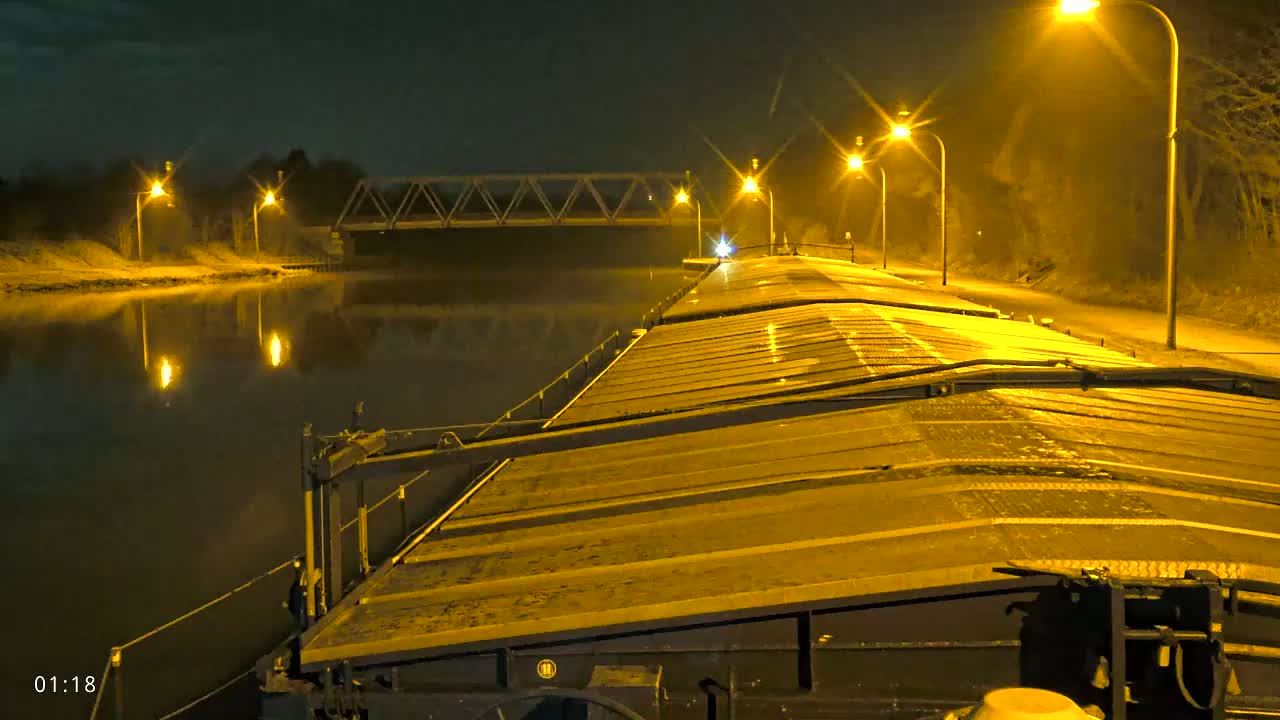 At night, a barge travels down a canal lit by streetlights, passing under a bridge.