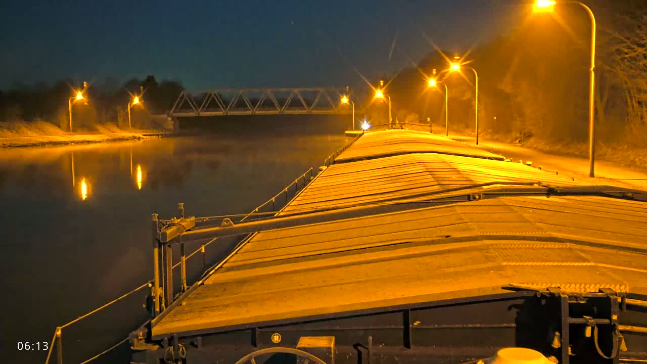 A barge illuminated by streetlights travels at night along a canal toward a bridge under a clear, dark sky.