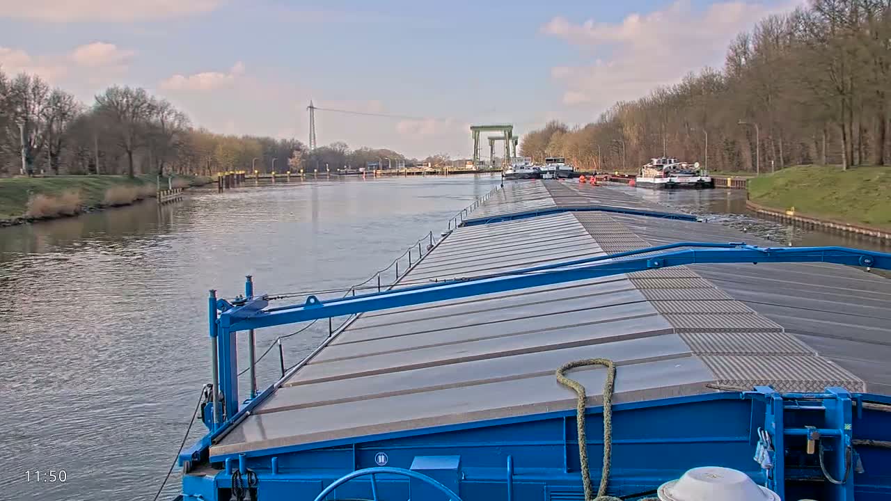 A blue barge travels down a calm canal under a partly cloudy sky, passing other barges near a lock.