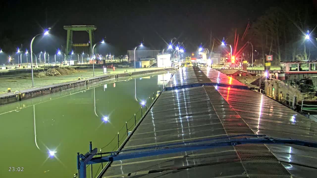 At night, a rain-slicked barge's deck is illuminated by lights reflecting off the calm, greenish water of a canal lined with more lights and docked vessels.