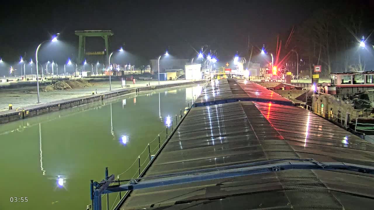 A nighttime view from atop a barge shows a calm, green canal lined with lights, alongside docked vessels and a crane.