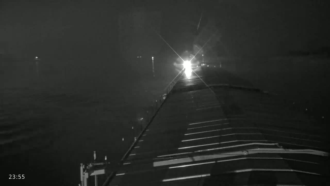 A nighttime view from the deck of a ship shows a bright light ahead on the water under dark, possibly stormy conditions.