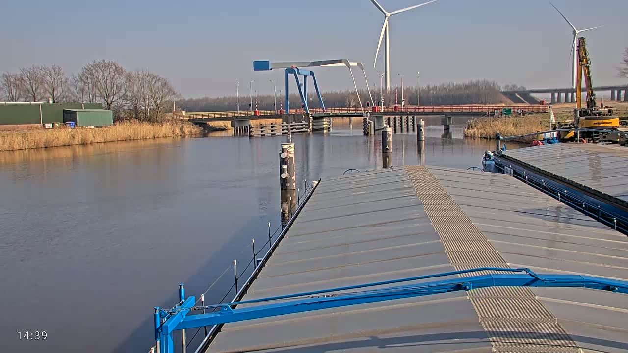 A barge on a calm waterway approaches a blue bascule bridge under a sunny sky, with wind turbines and construction equipment visible in the background.
