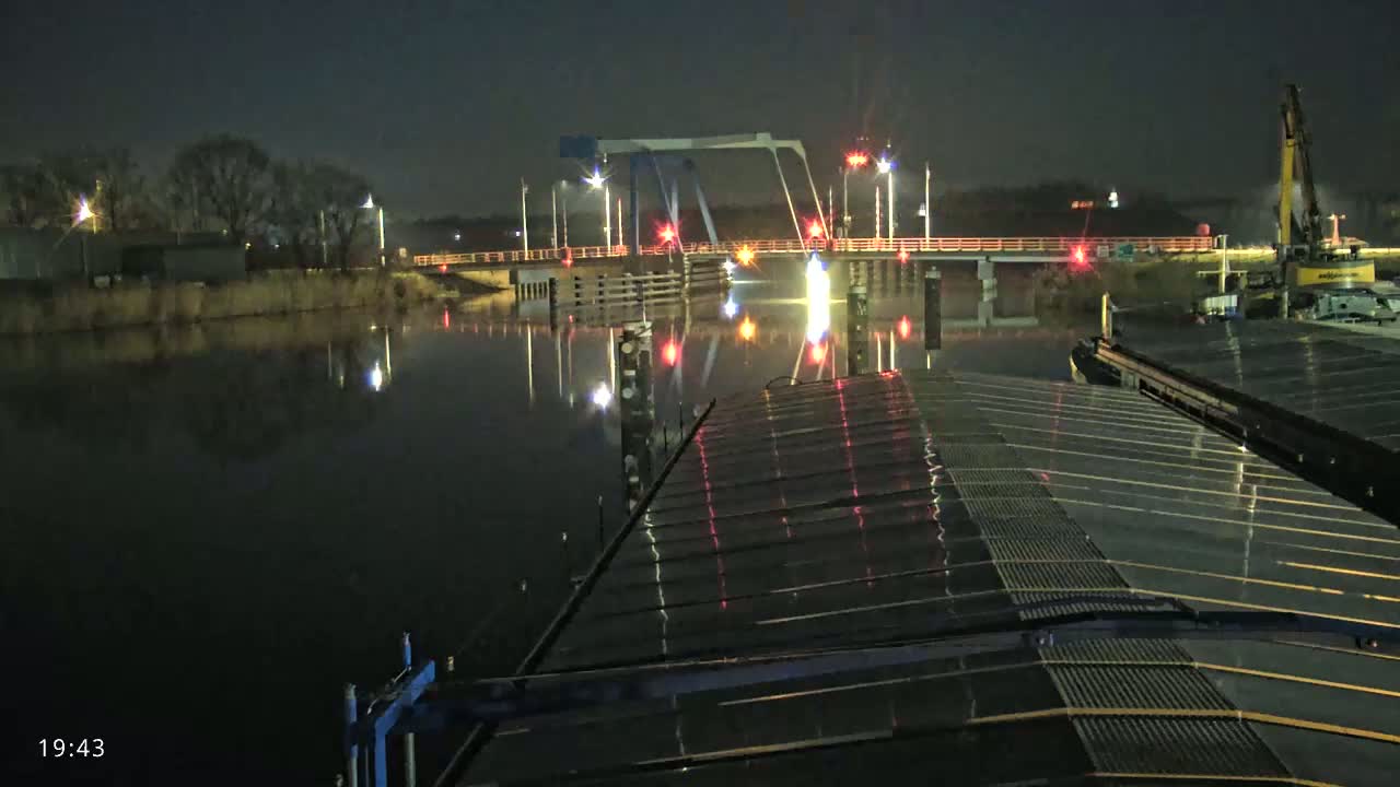 A nighttime view from a barge shows a lit-up drawbridge spanning a calm, dark river under a clear night sky.