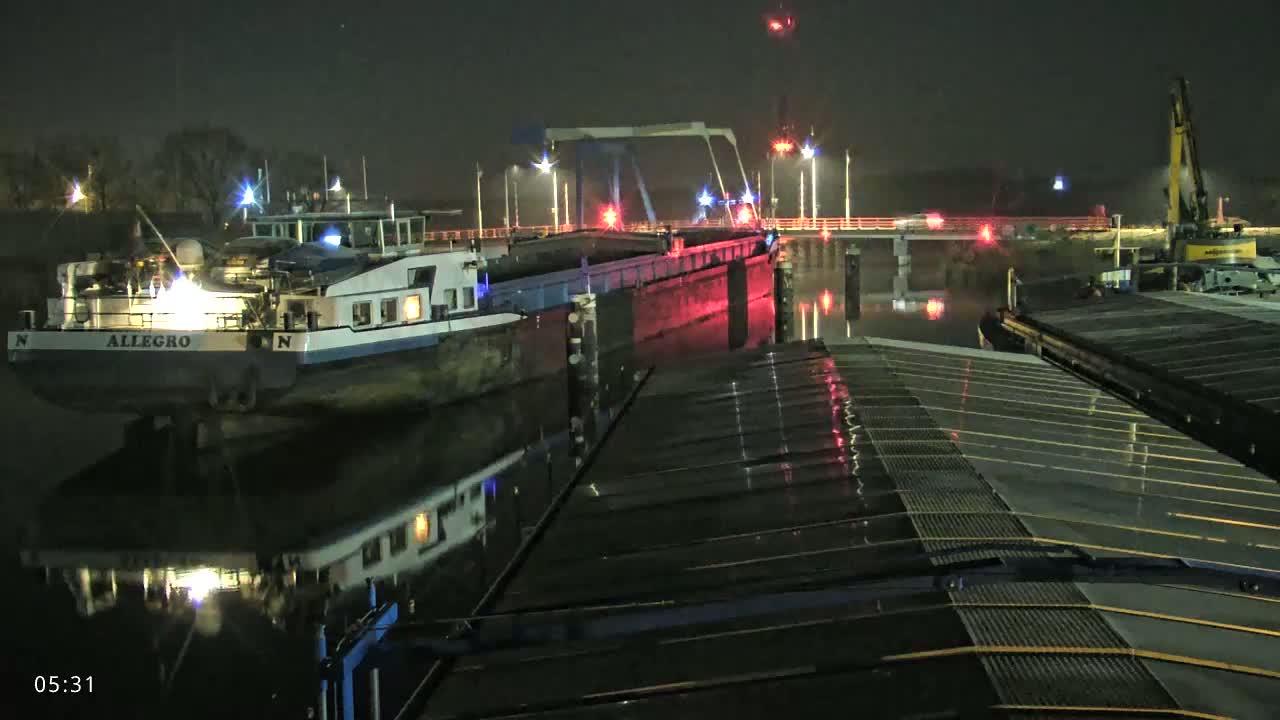 At night, a barge named Allegro is moored near a bridge under which another barge is passing, with a construction crane visible in the background.
