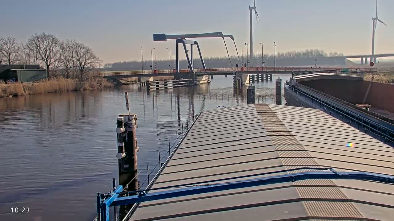 A barge approaches a lift bridge on a calm, sunny day, with wind turbines visible in the distance.
