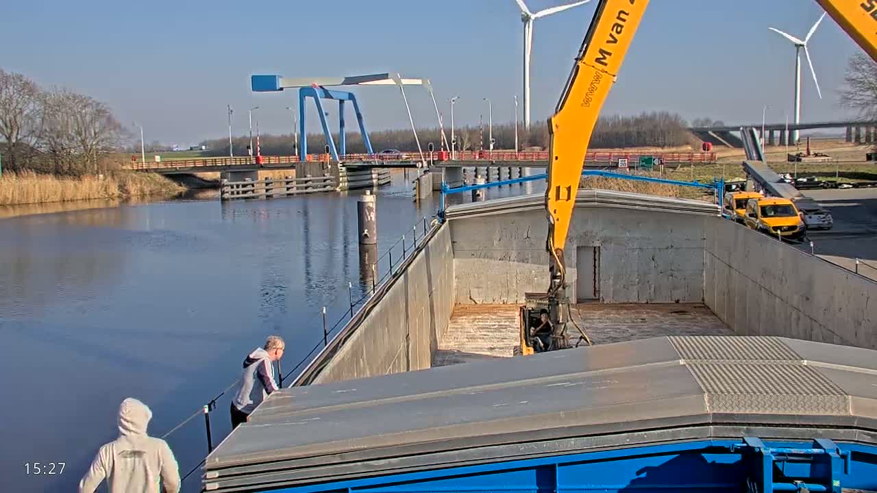 A large barge on a waterway is being loaded by a crane on a sunny day, with a blue drawbridge and wind turbines visible in the background.
