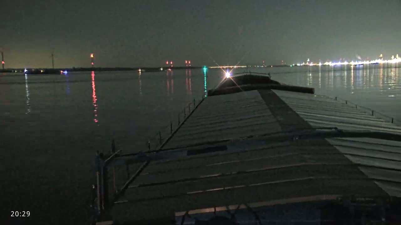A nighttime view from the deck of a barge on calm water, showing a distant city skyline illuminated with lights.