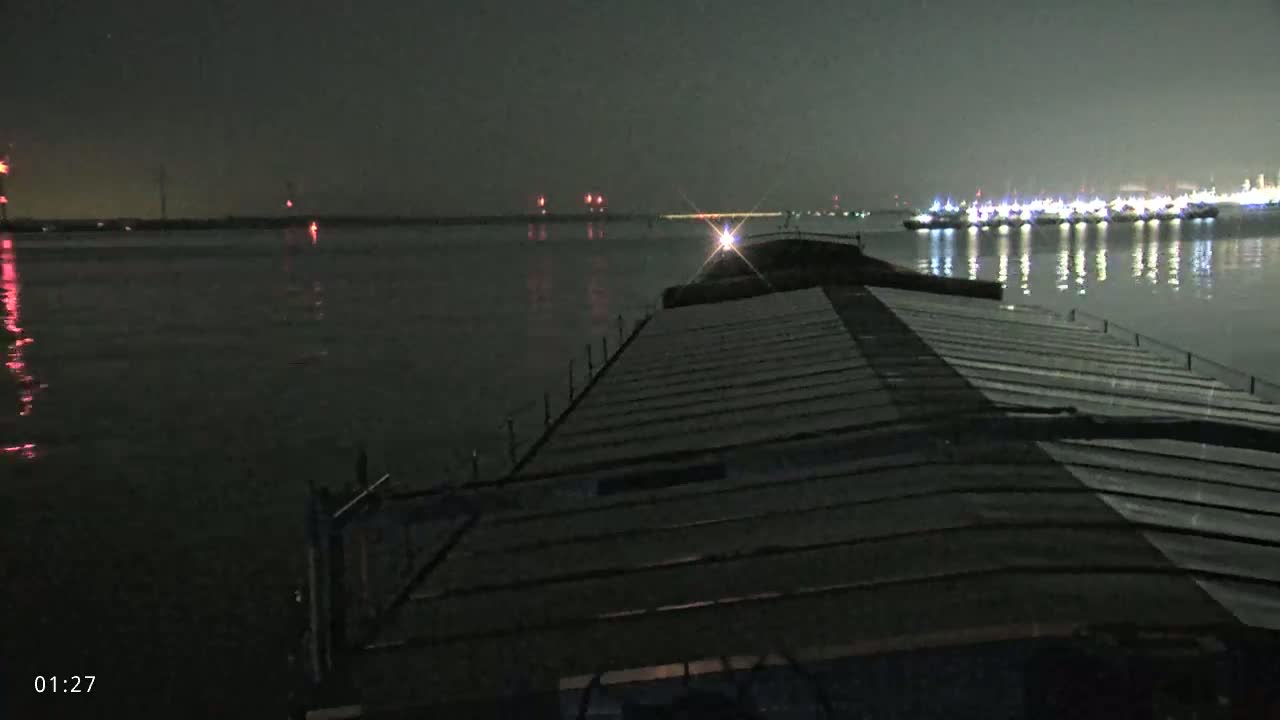 At night, from the deck of a barge, numerous lights are visible on the water's surface in the distance.