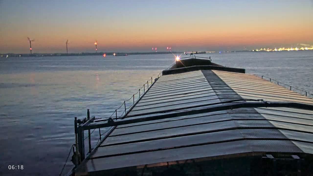 A barge travels on a calm body of water at sunset, with wind turbines and industrial structures visible on the horizon.
