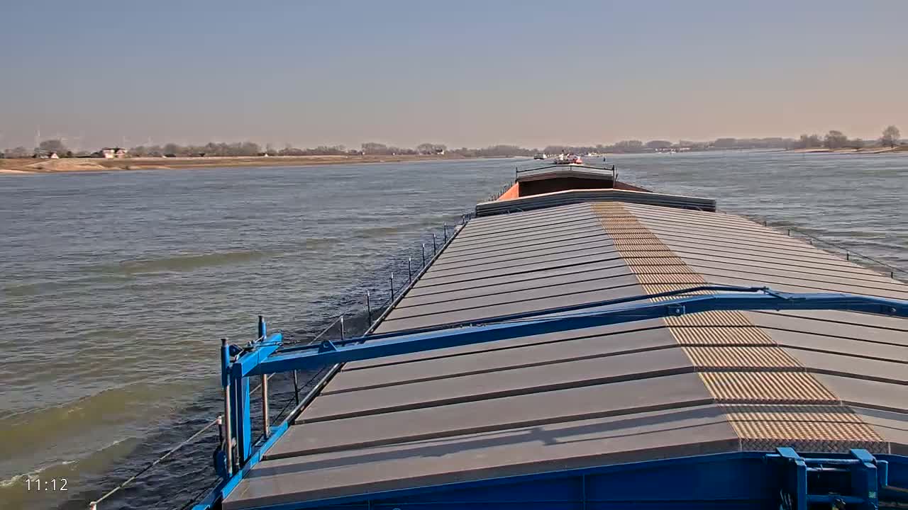 A barge travels down a wide river on a sunny day, other vessels visible in the distance.