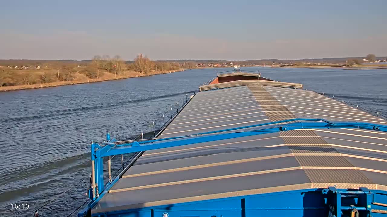 A barge travels down a wide river on a sunny day, with low-lying land visible on either side.