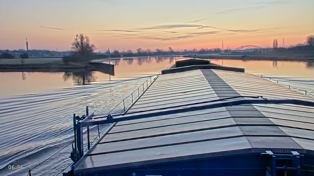 A barge travels down a wide river at sunrise, the calm water reflecting a pastel sky.