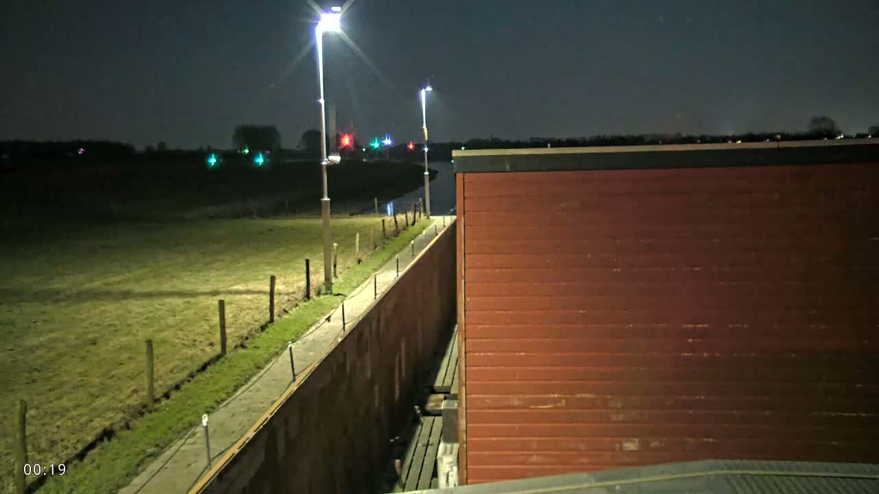 At night, a reddish-brown building is adjacent to a long, low wall beside a grassy field, lit by tall lampposts under a dark sky.