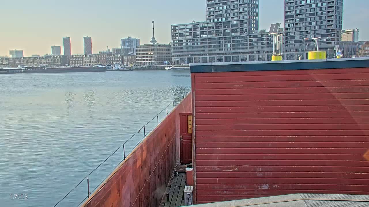 A red structure on a barge overlooks calm water, with a city skyline visible in the background under a clear sky.