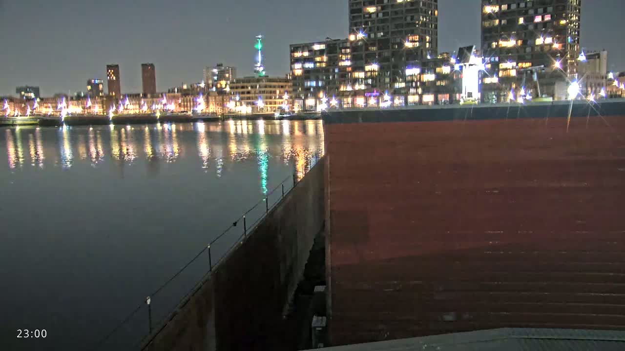A nighttime view of a calm body of water reflecting the lights of a city skyline and a large, dark-red vessel.