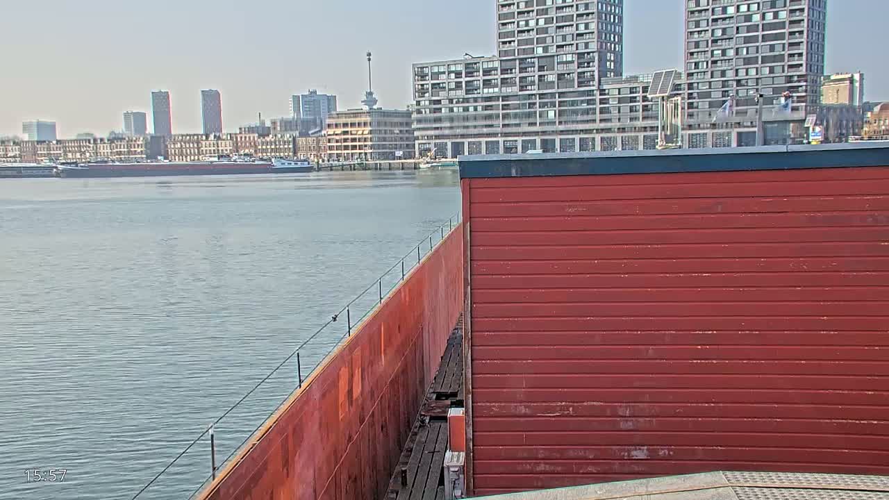 A red structure sits beside a body of water on a sunny day, with a cityscape of modern and older buildings in the background.