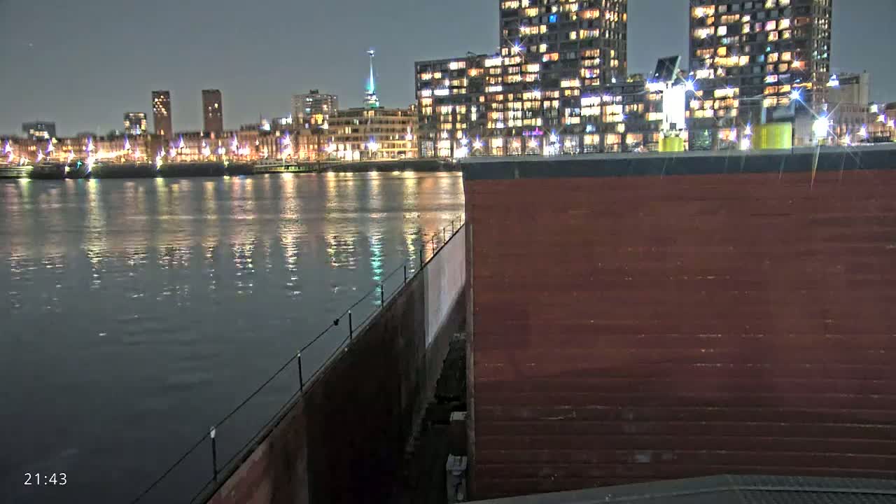 A nighttime view of a calm body of water reflecting illuminated city buildings, seen from the corner of a dark red structure.