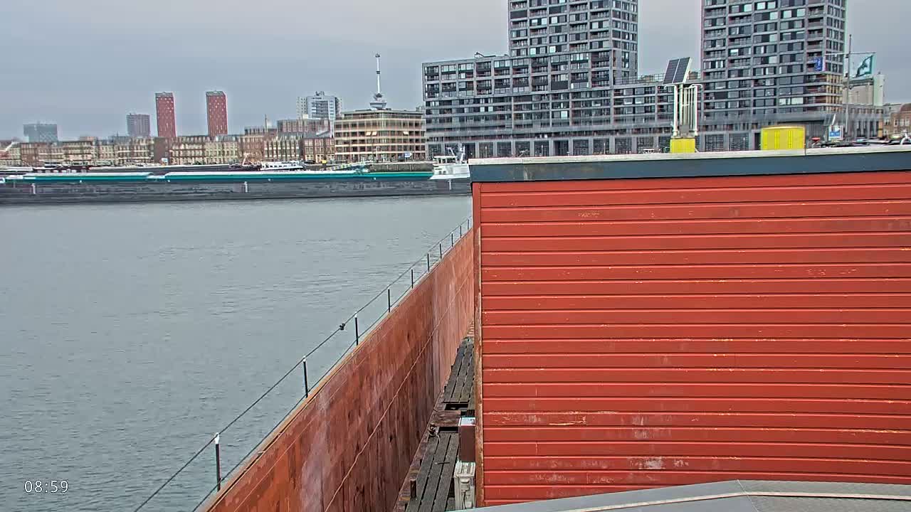 A rusty red barge is partially visible next to a bright red building, overlooking a waterway with a barge and a city skyline under an overcast sky.