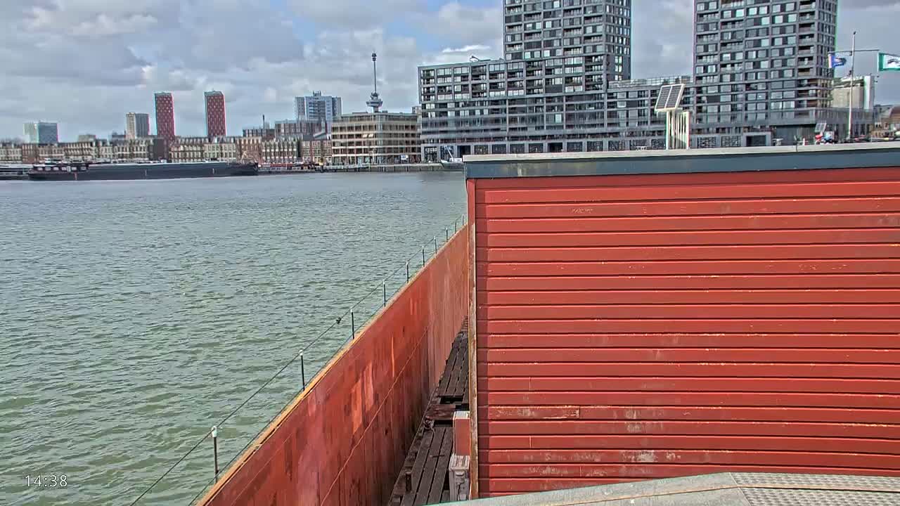 A red structure with horizontal siding sits next to a rusty brown barge, overlooking a calm body of water with a cityscape under a partly cloudy sky.