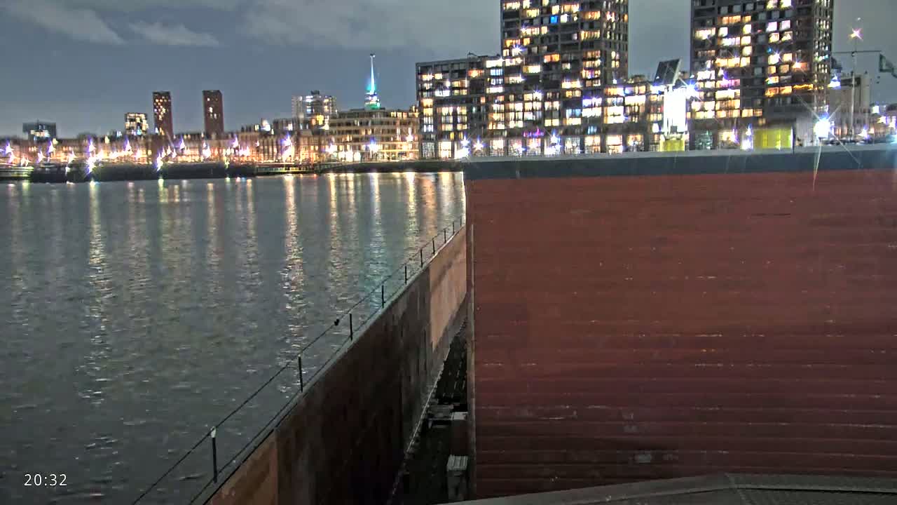 A dark-red structure, possibly part of a dock or lock, sits beside a calm body of water reflecting the lights of a city skyline at night.