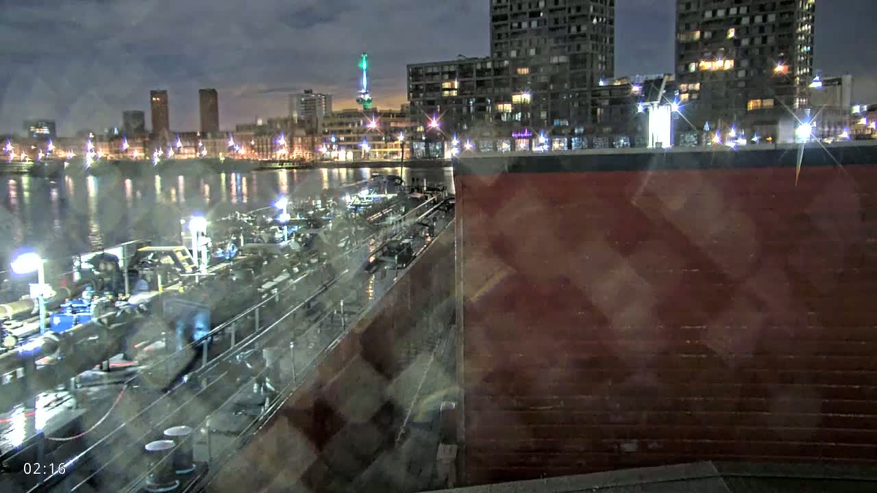 A long barge is docked at night next to a dark red wall, with a city skyline illuminated by lights across a calm body of water in the background.