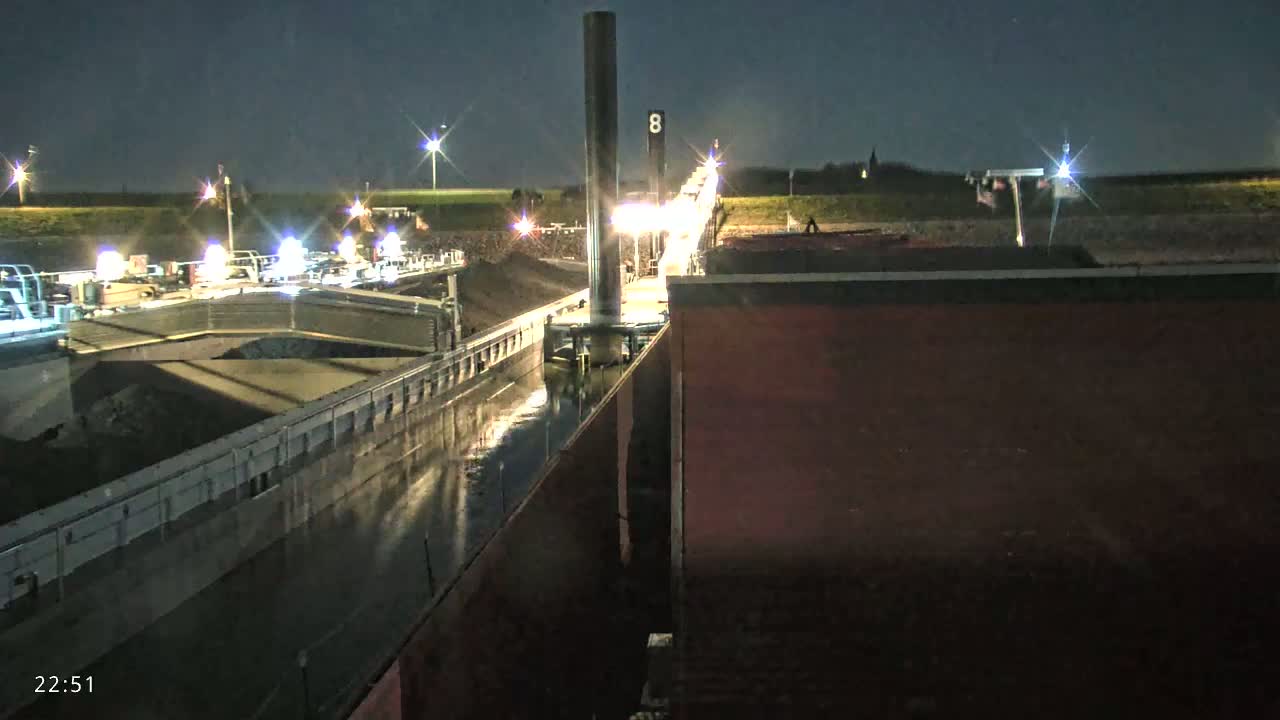 A barge carrying a dark cargo is illuminated at night beside a tall structure in a lock, under a clear, dark sky.