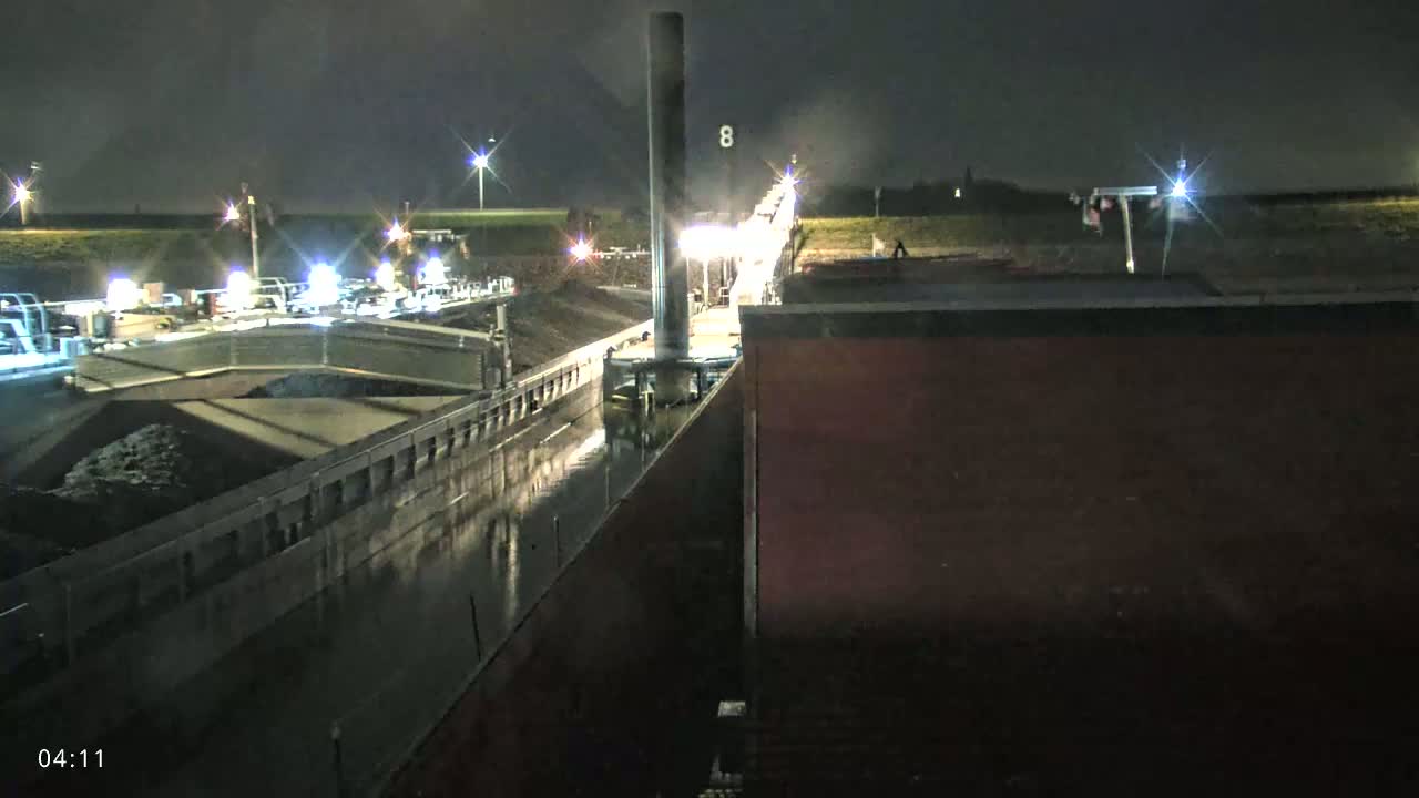At night, a barge loaded with dark material is in a lock, with other vessels and lights visible in the distance.