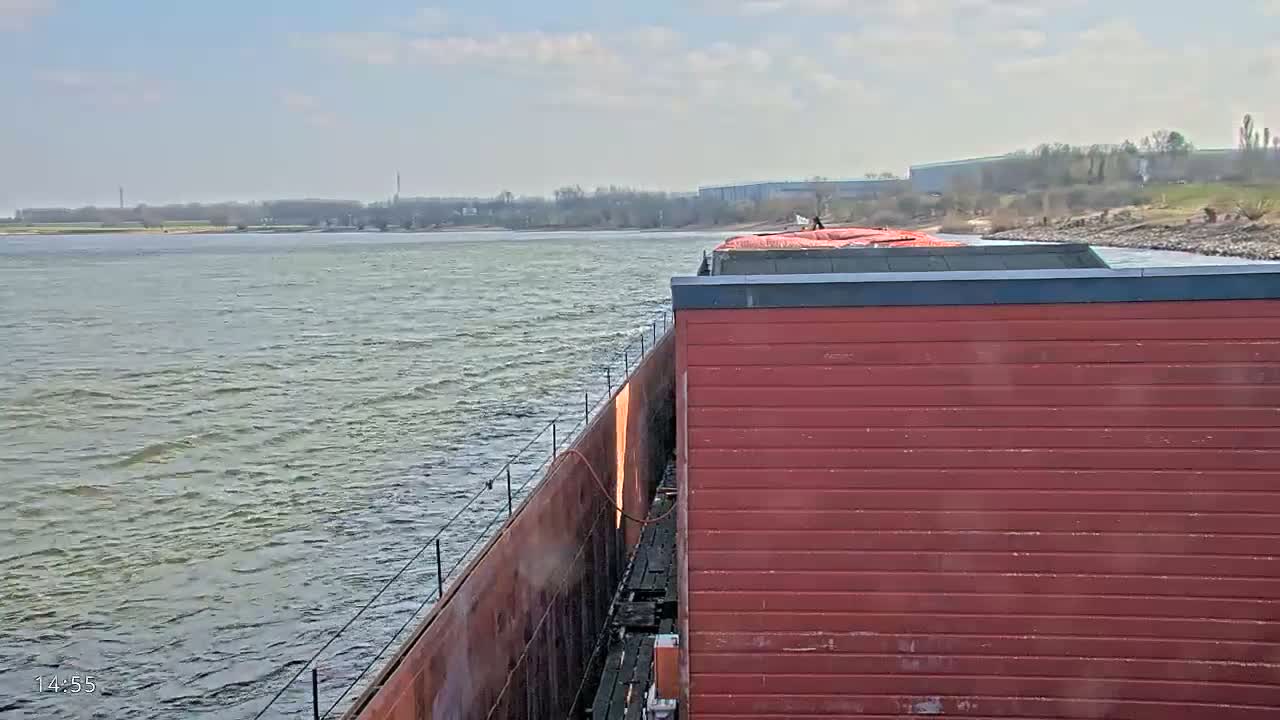 A red barge travels on a somewhat choppy river under a mostly clear sky.