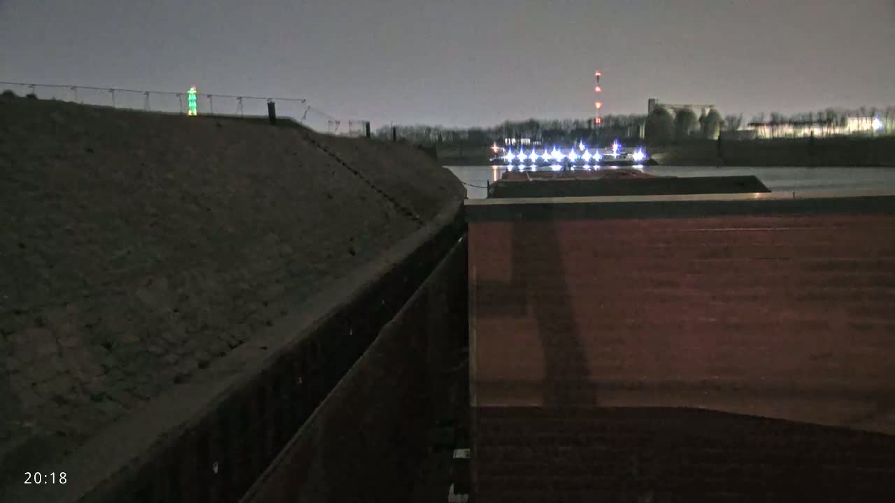 At night, a barge with lights is seen on a dark body of water next to a sloped embankment.