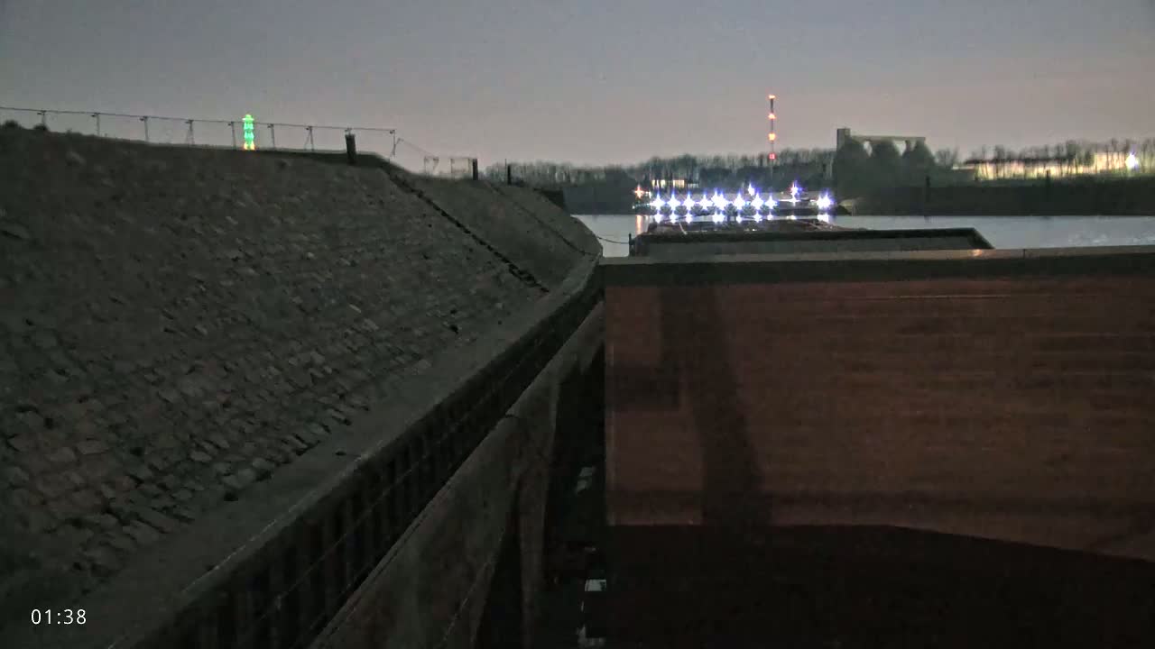 A nighttime view shows a dark, stone barge next to a lighted barge on a calm body of water.
