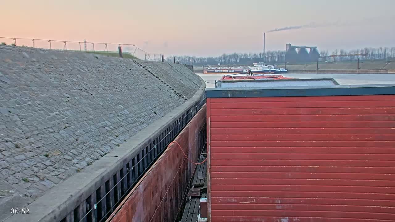 A red barge is moored next to a stone embankment, with other barges and industrial structures visible in the distance under a pale sky.