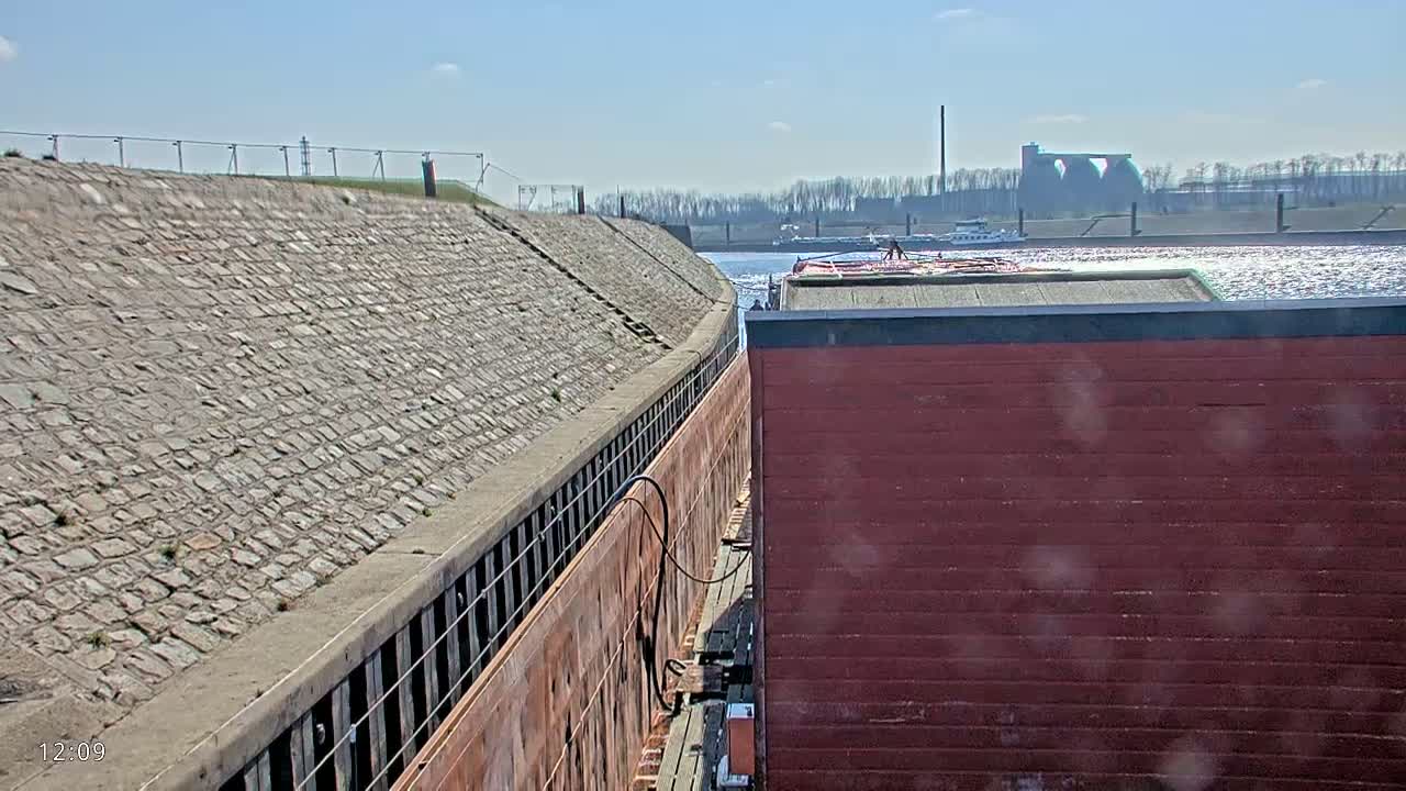 A red barge is docked next to a stone embankment on a sunny day, with a larger barge visible in the distance on calm water.