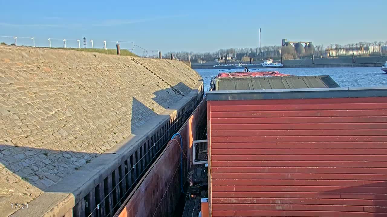 A red barge is docked next to a stone retaining wall on a sunny day, with other barges and vessels visible in the distance on calm water.