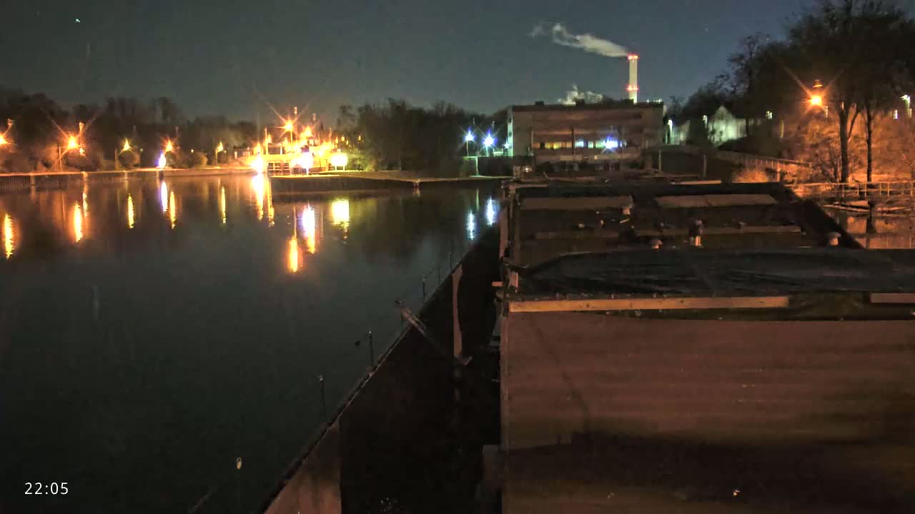 A nighttime view of a calm body of water reflecting lights from a nearby industrial area and shoreline, under a clear, dark sky.