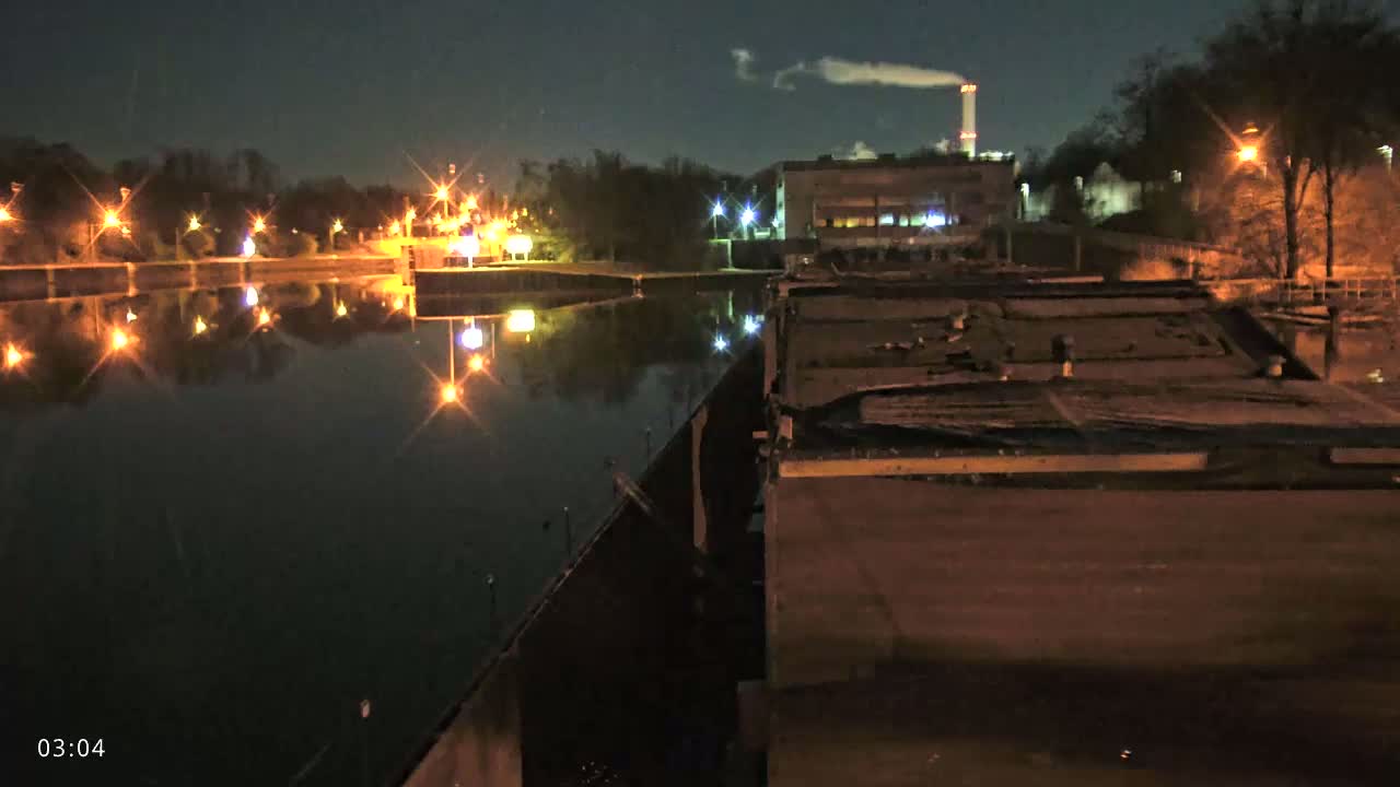At night, a calm body of water reflects the lights of a shoreline and a nearby industrial building emitting smoke.