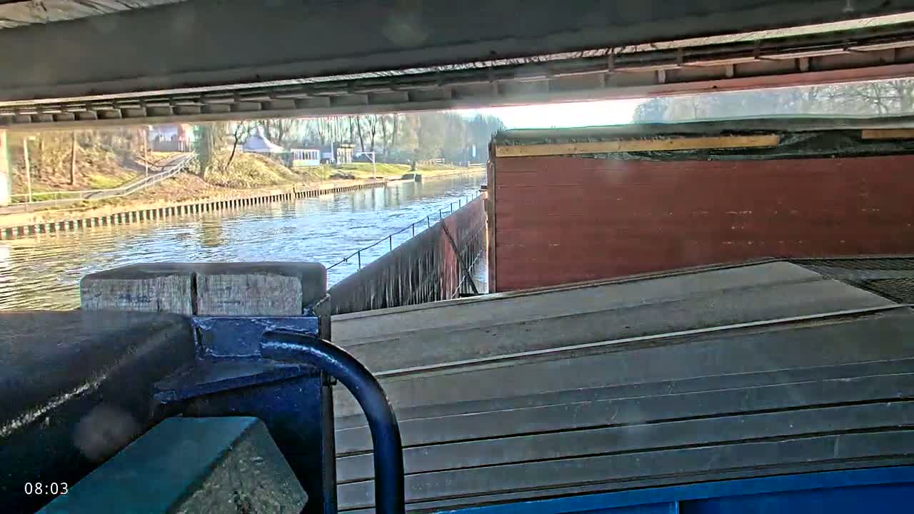 A view from a barge on a calm, sunny day, passing under a bridge and alongside a canal bank.