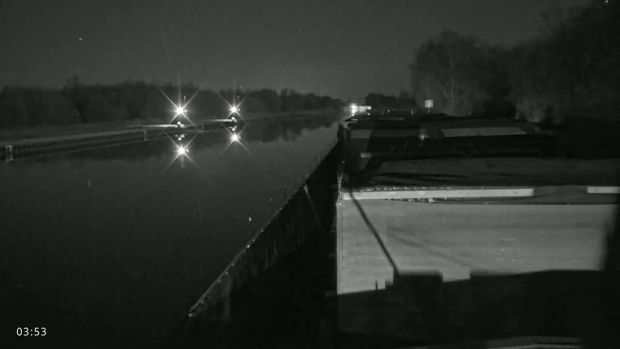 A nighttime view from a barge on a calm canal shows distant headlights approaching and trees lining the banks.