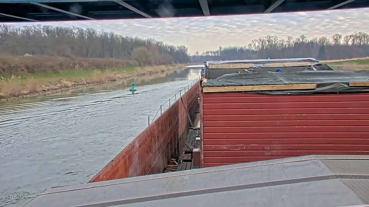 A barge travels down a calm river under a cloudy sky, passing by bare trees lining the riverbank.
