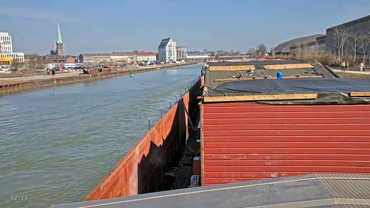 A red barge travels down a canal on a sunny day, passing a cityscape with buildings and a church steeple visible in the distance.