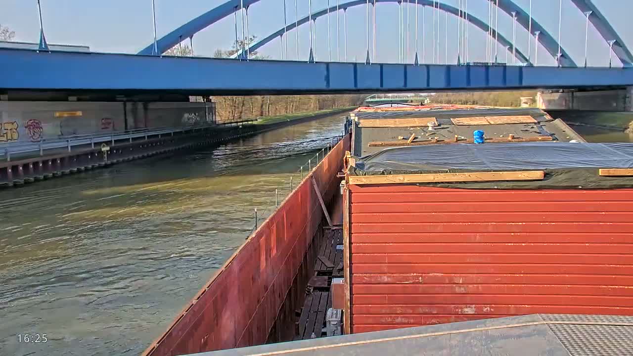A red barge travels on a canal under a blue arched bridge on a sunny day.